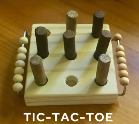 Bohrmaschinenführerschein: Holzspielzeug mit sieben vertikalen Stäben auf einem quadratischen Brett mit Griffen und Perlen an den Seiten, auf einem Holztisch. Text "TIC-TAC-TOE" darunter.